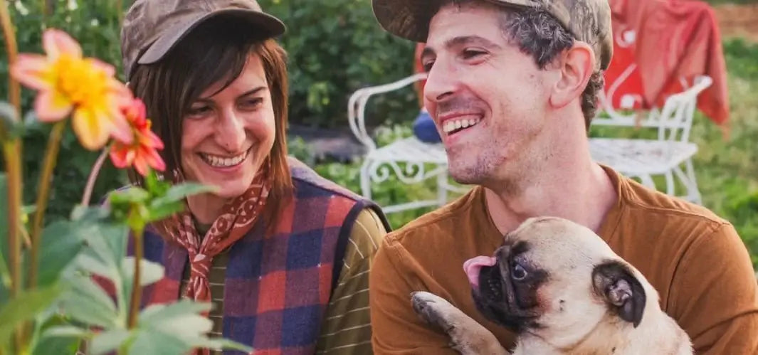 Two people, one holding a pug, standing in a garden with flowers and chairs in the background.