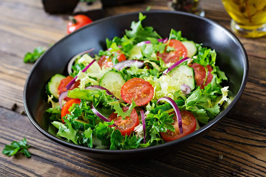 Fresh salad with tomatoes, cucumbers, and greens in a black bowl on a wooden table.